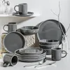 A collection of grey stoneware dishes and mugs with white trim, arranged on a surface with a light gray background. Some items are stacked, while others are placed individually. Náhled obrázku produktu