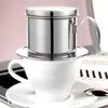 A stainless steel Vietnamese coffee filter sits atop a white coffee cup, which is placed on a saucer. A laptop and some papers are visible in the background. Produktbild-Vorschau