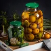 Two jars of preserved food, one with apples and the other with pickles and garlic, sit on a wooden tray with some fresh produce and herbs. Podgląd zdjęcia produktu