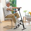 An elderly man is using a pedal exerciser while sitting on a chair in a living room. Produktbild-Vorschau