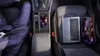 A person's legs are visible on the left side of the image, with a laptop and tablet charging on the car seat on the right. The car's center console is visible, with a phone charging wirelessly. Anteprima dell’immagine prodotto