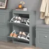 A gray shoe cabinet with two flip-down compartments filled with shoes, next to a gray drawer unit and a gray bench with a gray cushion. A trench coat is hanging on the wall to the right. Produktbild-Vorschau