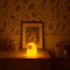 A cute, glowing bear-shaped night light sits on a wooden table next to two suitcases, with a framed picture on the wall behind it. The room has a warm, inviting ambiance. Náhľad obrázku produktu