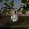 A solar-powered light bulb shaped lantern hangs from a tree branch at dusk. Produktbild-Vorschau