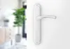 A close-up of a silver door handle on a white door, with a blurred interior scene in the background. Produktbild-Vorschau