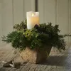 A white LED candle is placed in a square planter filled with greenery, pinecones, and a small dried flower. The planter sits on a rustic wooden table, with a few star-shaped ornaments in the foreground. Produktbild-Vorschau