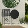 A black metal bistro set with a round table and a slatted chair is placed on a gravel patio next to a concrete wall with a circular opening revealing greenery. An aloe plant is situated to the right of the table. Produktbild-Vorschau