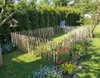A garden scene with a rustic wooden fence enclosing a grassy area, surrounded by flowers and trees. Produktbild-Vorschau