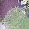 Two crocheted doilies, one purple and one green, with decorative edges, are arranged together with some flowers. Produktbild-Vorschau