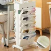A mobile magazine organizer with multiple shelves is placed next to a desk, with a person sitting at the desk. Produktbild-Vorschau