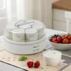 A yogurt maker with several jars filled with yogurt, placed on a table with a bowl of berries and a small bowl of yogurt. Produktbild-Vorschau