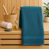 A teal towel is draped over a wooden rack in a sauna setting, with a wooden bucket, ladle, rolled white towel, a small bottle of oil, and a stone on a wooden bench. Produktbild-Vorschau