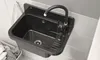 A black granite composite sink with a chrome faucet and soap dispenser is mounted on a tiled wall. A laundry basket with clothes is visible to the left. Produktbild-Vorschau