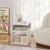 A white side table with two open shelves is placed next to a textured armchair, with a framed picture and a cup of coffee on top. Produktbild-Vorschau
