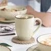 A close-up of a beige mug filled with a beverage, placed on a jute coaster on a white table. In the background, a bowl of food and a person's arm are visible. Náhled obrázku produktu