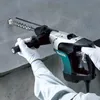 A person wearing white gloves is using a Makita hammer drill to drill into a concrete wall. Produktbild-Vorschau