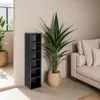 A tall, narrow black shelf unit stands next to a large potted plant and a beige sofa in a living room. Produktbild-Vorschau