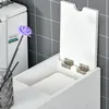 A close-up view of a white bathroom storage unit with its lid open, revealing paper products inside. A decorative purple flower in a vase is visible to the left. Voir les images du produit