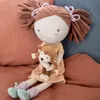 A plush doll sitting on a textured surface, holding a small monkey plush. Produktbild-Vorschau