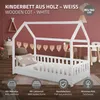 A white wooden house-shaped cot is shown in a child's bedroom setting with decorative elements and furniture in the background. Produktbild-Vorschau