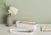 A white storage box filled with pencils, a tray with scissors, and a vase with a flower and grass on a white surface against a light green background. Produktbild-Vorschau