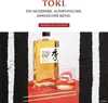A bottle of Suntory Toki Japanese whisky is displayed next to a glass of whisky with ice and chopsticks, set against a striking black and white striped background with a red surface. Produktbild-Vorschau