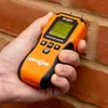 A hand holding an orange and black moisture meter against a brick wall. Voir les images du produit