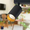 A close-up of a black ceiling spotlight with a gold interior, mounted on a track. The background is blurred, showing a domestic interior with furniture. Náhľad obrázku produktu
