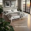 A white metal bed frame with bedding is set up in a bedroom with large windows and plants. Náhled obrázku produktu