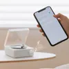 A hand holding a smartphone is shown interacting with a small, white, record-player-shaped Bluetooth speaker. Podgląd zdjęcia produktu