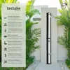 A solar shower is shown outdoors in a garden setting, surrounded by plants and a stone pathway. Náhled obrázku produktu