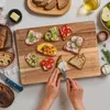 A close-up overhead shot of a wooden cutting board with various open-faced sandwiches and a person spreading cream cheese on a slice of bread. Produktbild-Vorschau