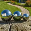 Three polished stainless steel garden spheres of different sizes are placed on a weathered wooden surface in a garden setting. A paved pathway winds through a manicured lawn with flower beds in the background. Produktbild-Vorschau