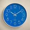 A blue wall clock with white numbers and hands is mounted on a textured light gray wall. Náhled obrázku produktu