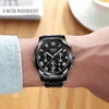 A man's wrist wearing a black watch with a black dial and silver accents. The background is blurred and shows a wooden surface and a white cup. Produktbild-Vorschau