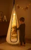 A young boy is standing next to a hanging cocoon chair in a dimly lit room, holding a teddy bear. Produktbild-Vorschau