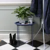 A navy blue side table with a plant and an oil burner on it, placed on a black and white checkered floor in front of a light gray wall with wainscoting. Two black boots are on the floor next to the table, and a coat rack with navy blue coats is to the righ Produktbild-Vorschau