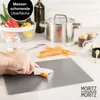 A person is chopping carrots on a cutting board, with kitchen items like a pot, vegetables, and spices in the background. Produktbild-Vorschau