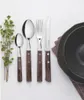 A set of cutlery with wooden handles is arranged on a white surface next to a sprig of greenery and a dark bowl. Podgląd zdjęcia produktu