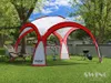 A red and white event pavilion set up on a green lawn with trees and a wall in the background. A lounge chair and a grill are also present. Produktbild-Vorschau