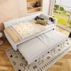 A white daybed with a pull-out trundle bed is set up in a room with a window overlooking a green landscape. Pillows and bedding are arranged on the daybed. Náhled obrázku produktu