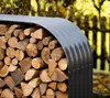 A close-up shot of a metal firewood storage rack filled with logs, set outdoors with a blurred background of trees and a fence. Náhled obrázku produktu