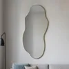 A large, organically shaped mirror with a gold frame hangs on a light gray wall above a sofa. A floor lamp is visible to the left of the mirror. Náhľad obrázku produktu