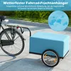 A bicycle with a blue cargo trailer attached, parked on a paved surface with greenery in the background. A circular graphic showing water droplets is in the upper right corner. Produktbild-Vorschau
