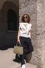 A person stands leaning against a stone wall, holding a handbag. Voir les images du produit