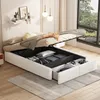 A bedroom scene with a white upholstered bed frame, its storage compartment open, revealing organized items. The bed is made with neutral-toned bedding and a throw blanket. A large window with a view of greenery is visible in the background. Produktbild-Vorschau