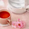 A cup of tea and a glass kettle are placed on a pink tablecloth, with pink flowers nearby. Produktbild-Vorschau