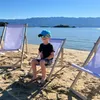 A young boy sits on a beach chair on a sandy beach, with the ocean and mountains in the background. Podgląd zdjęcia produktu