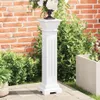 A white pedestal stand with a floral arrangement on top, placed outdoors on a patio next to green bushes and a brick wall. Anteprima dell’immagine prodotto