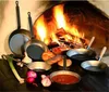 A collection of cast iron pans arranged in front of a roaring fireplace, with some ingredients like onions, garlic, and bread scattered around. Anteprima dell’immagine prodotto